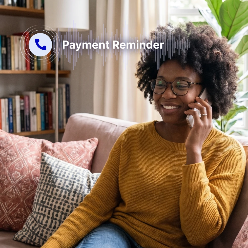 Smiling woman wearing mustard-yellow ribbed sweater and glasses, sitting on beige couch with patterned pillows in bright living room. Bookshelf and green plant visible in background with natural window light. Woman holds phone to ear during call. Overlaid phone icon with sound wave graphic and white text reading "Payment Reminder."